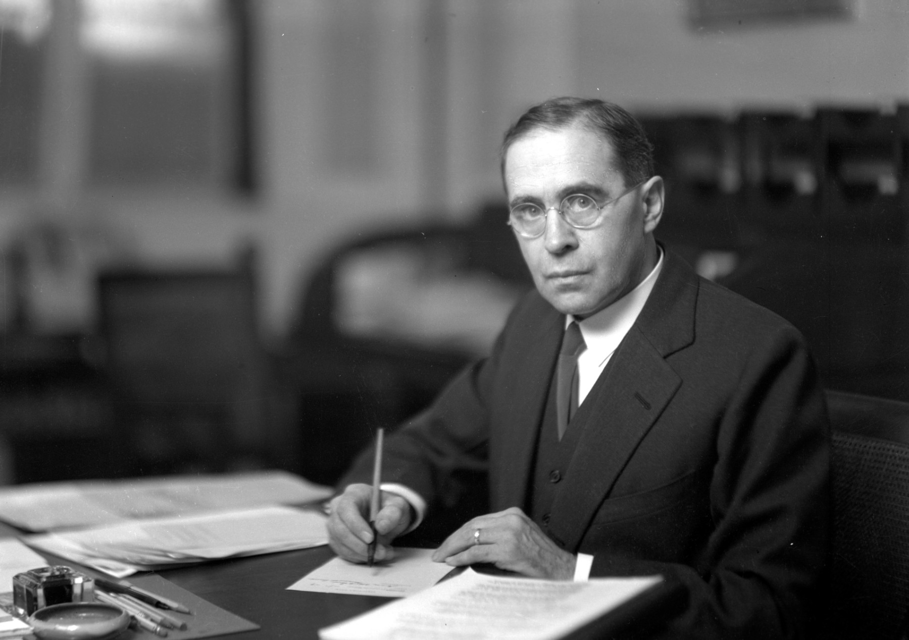 A black-and-white photograph of a man sitting at a desk with a writing implement in hand. He wears a dark formal suit and has round glasses.