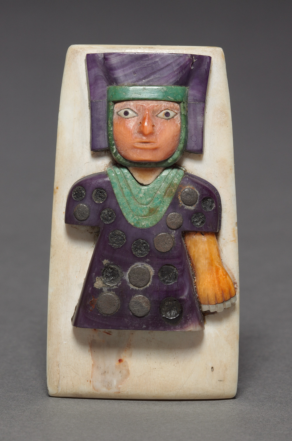 A small ornament that has a white shell base with a figure attached to the front. The figure is depicted from the waist up with one arm and dressed in dark purple attire and headdress. Their necklace is greenstone and round metal elements have been embedded into their torso.