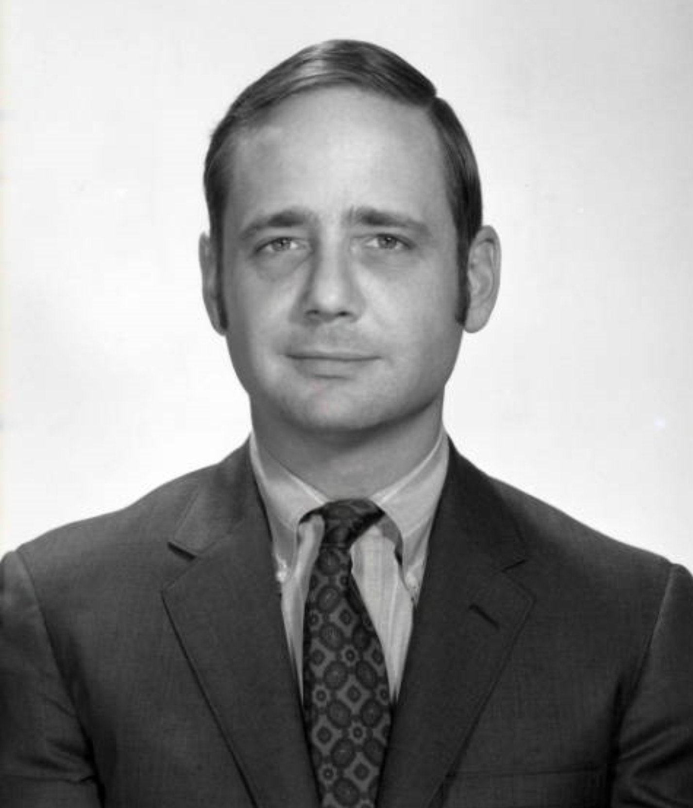 A black-and-white photograph of a man, from the bust up, dressed in a formal suit with tie. He is clean shaven and looks directly into the camera.