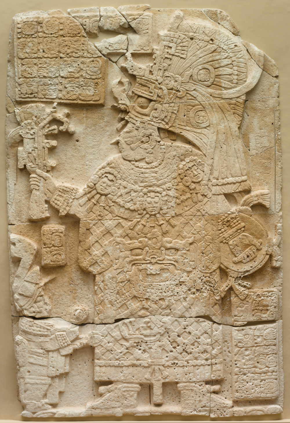 A light tan stone sculpture depicting a woman dressed in elaborate regalia holding a weapon and shield. Her body faces forward but her head is turned in profile. A smaller figure stands next to her beneath the hand wielding the weapon. Panels of glyphic writing surround her.