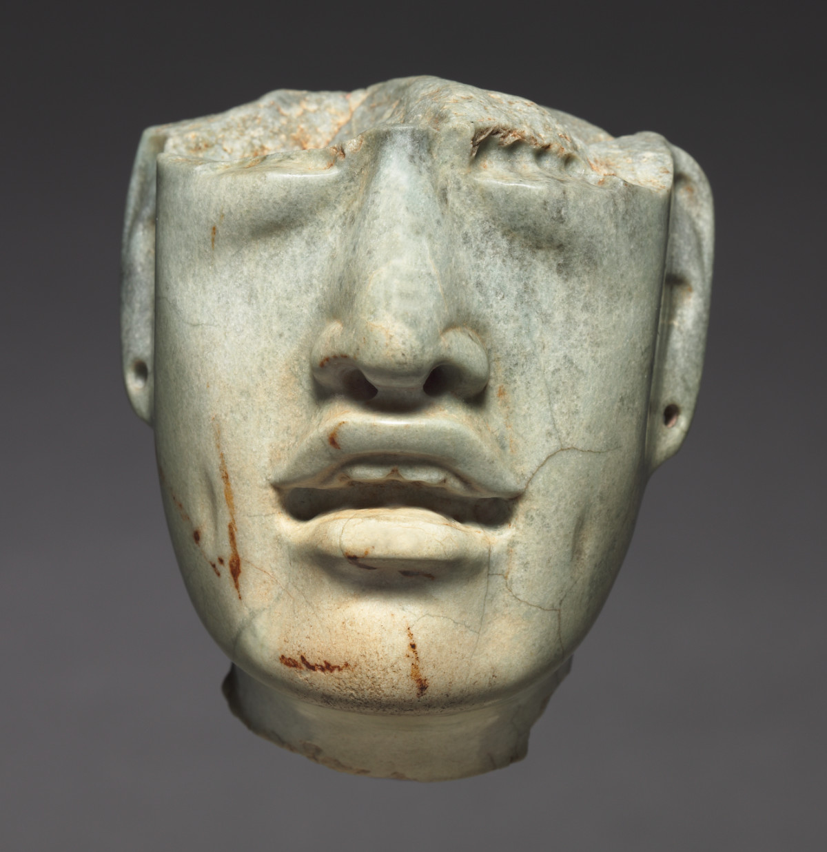 A greenstone sculpture of a head and partial neck. The stone above the cheekbones is broken, leaving the face without eyes. The earlobes have holes drilled in, and the mouth is carved to be slightly open.