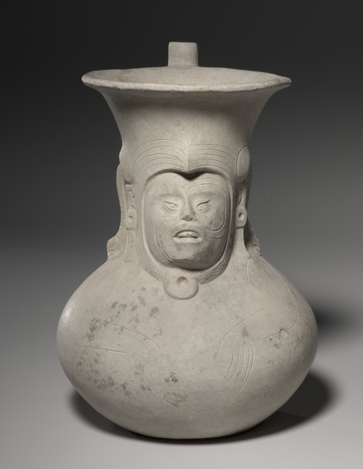 A white-colored ceramic depicting a face in the neck of the jar. The facial features and headdress are relief carved while the arms are lightly incised on the body of vessel.