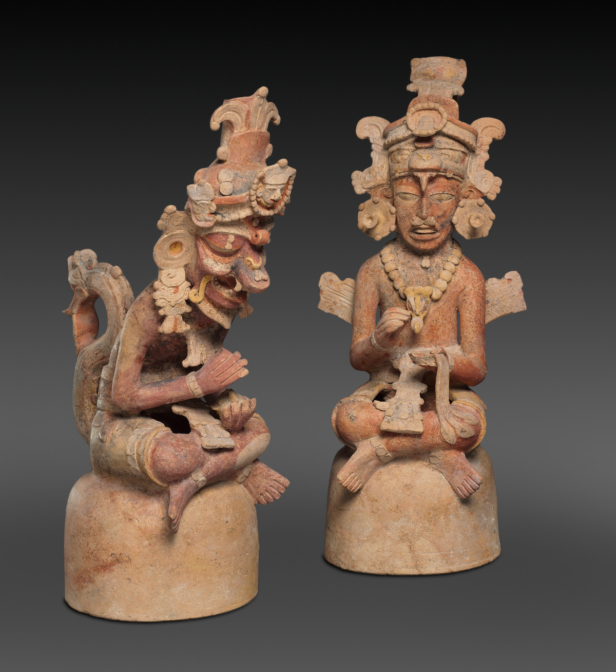 Two figures seated cross legged on round stools, leaning forward with empty hands that once would have held implements for artwork. They wear elaborate headdresses, clothing, and accessories.