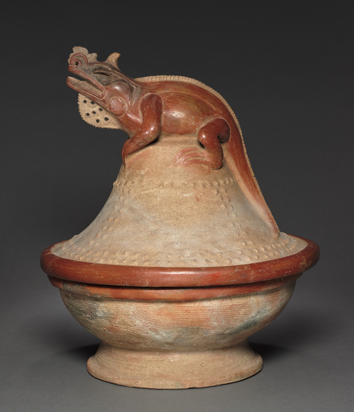 A tan ceramic bowl with a bell-shaped lid that is wider on the bottom and narrower at the top and that bears an iguana on top. The iguana is dark red with its head tilted up and minimal decorative features. The tail follows the side of the lid.