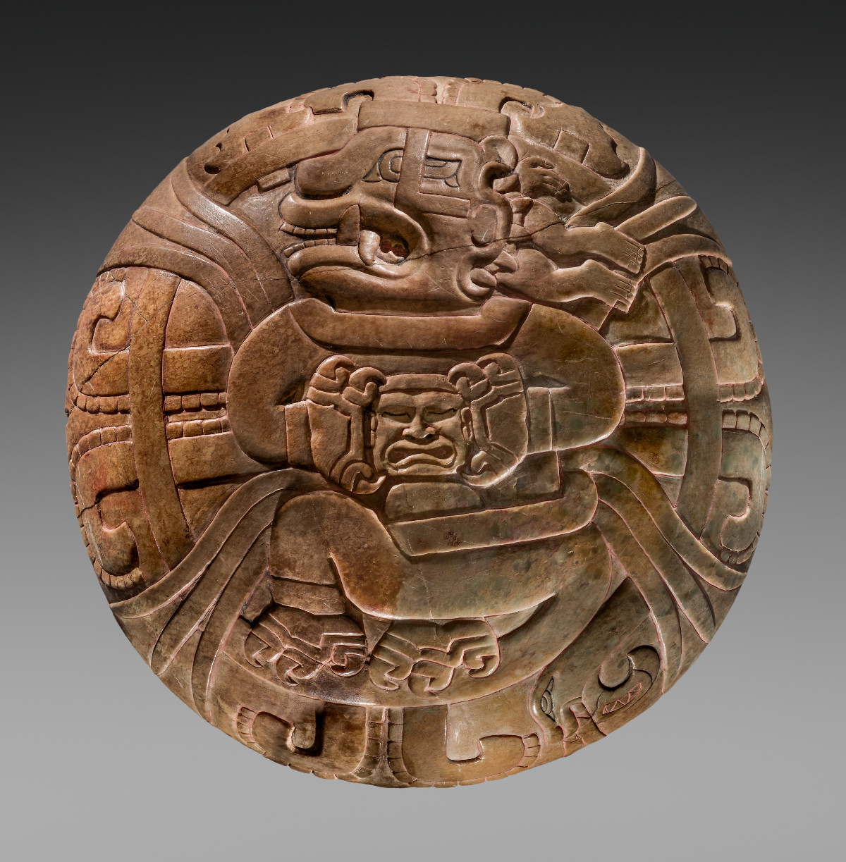 A round, brown plate with a supernatural figure curled in the center holding a face to its chest. The supernatural figure bares its teeth, and a pair of legs unattached to a body can be seen behind it. The rest of the plate is highly decorated.