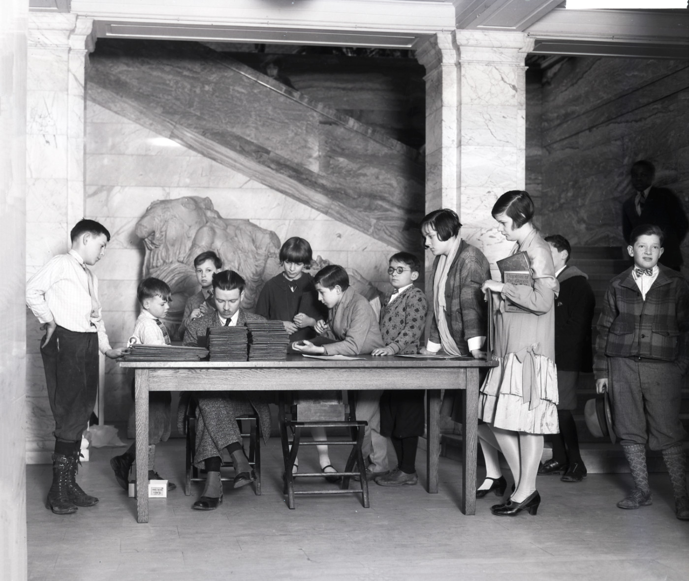 A black-and-white photograph of a man seated at a table in a marble foyer. A large group of children stand next to him and peer over his shoulder.