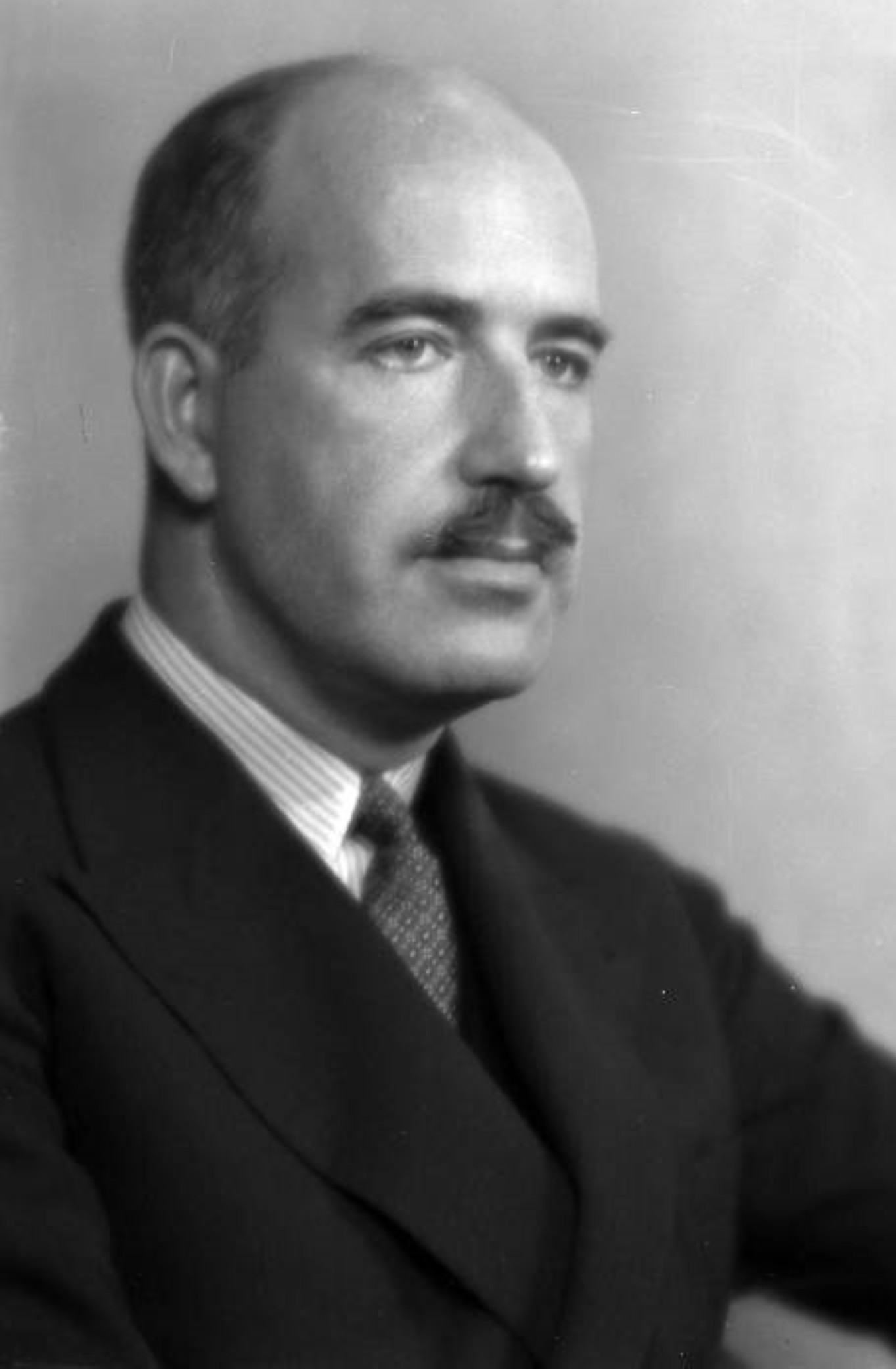 A black-and-white photograph of a bald middle-aged man turned three-quarters away from the camera. He is wearing a formal suit and has a mustache.
