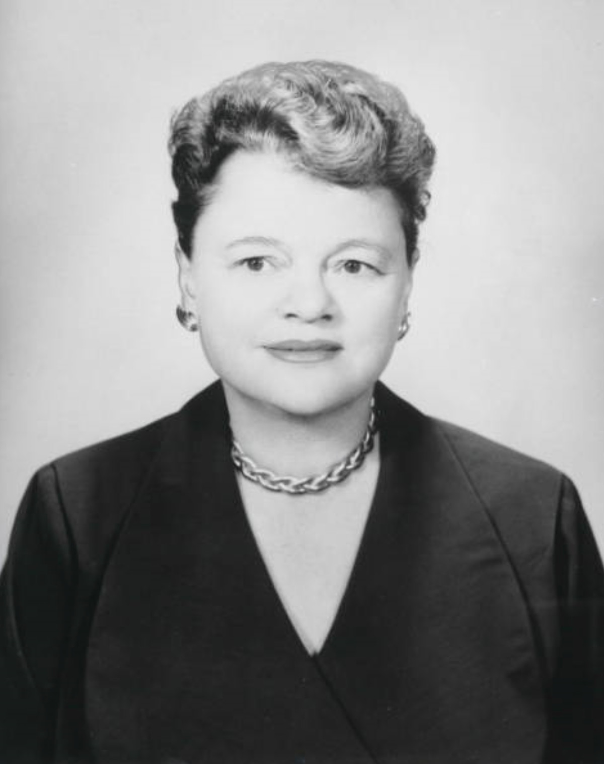 A black-and-white photograph of a woman from the bust up, facing the camera. She wears a dark suit jacket, a thick chain necklace, large earrings, and her hair is cut short.