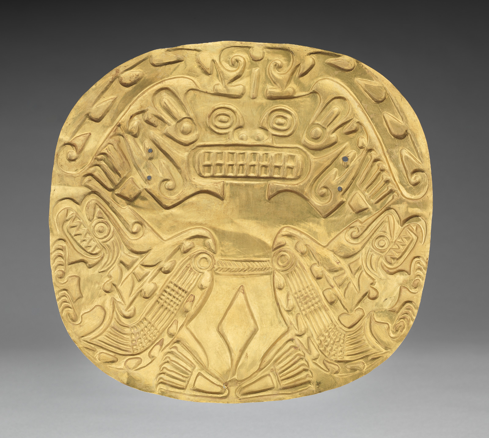 A round gold plate that would be hung from the four small holes in the center. It depicts a highly decorative figure with prominent teeth. Two small creatures stand on either side of the central figure.