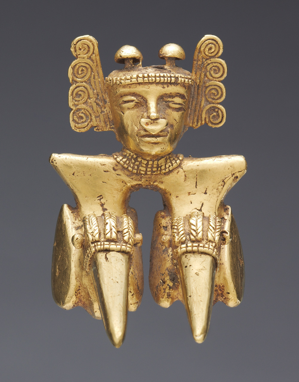 A small golden bust sculpture of a figure with an elaborate headdress, septum nose piercing, and collar necklace. At the base of the shoulders two protrusions extend into points with light decorations carved into them.
