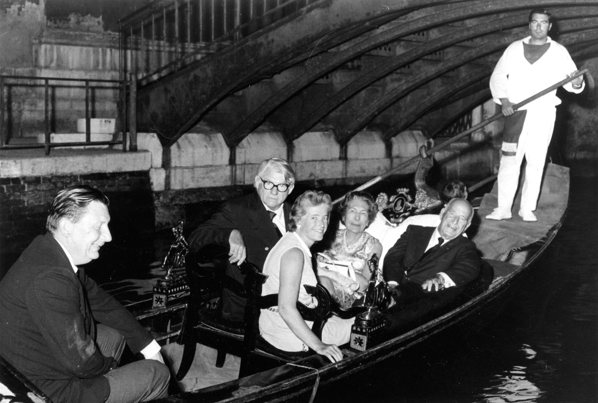 A black-and-white photograph of five people sitting on a gondola in a river while a man dressed in all white rows. The people seated in decorative carved chairs wear formal clothing and are turned to pose for the camera.