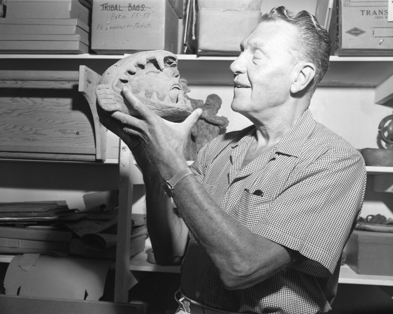 A black-and-white photograph of a man holding a ceramic anthropomorphic shaped figurine close to his face, looking tenderly at the object. He is dressed casually and stands in front of shelves of boxes.