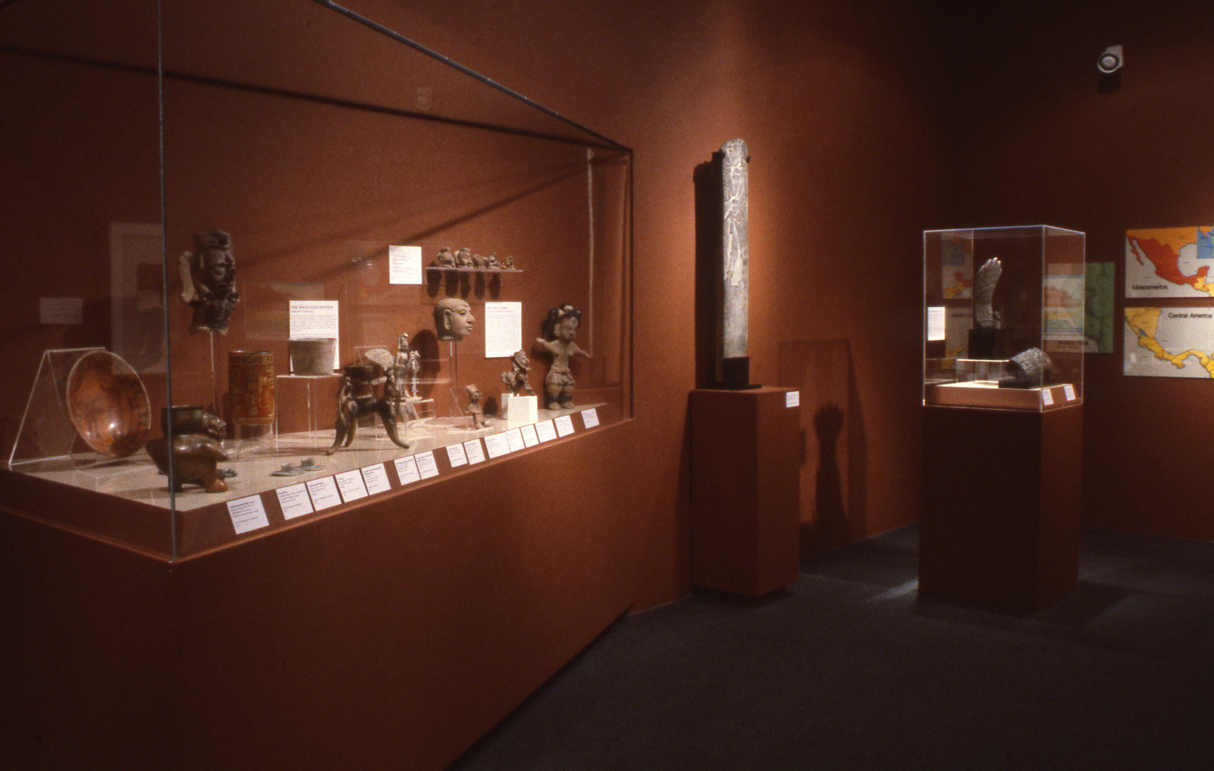 A gallery installation photograph of the pre-Columbian collection. There are two filled cases and one pedestal that displays the Veracruz stone slab. The gallery space is painted dark orange, and on the back wall are several maps of Mesoamerica and Central America.
