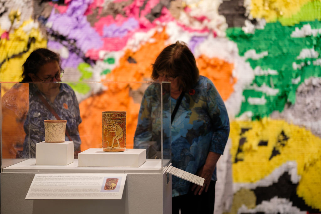 A photograph of two women standing in the exhibition space reading a label attached to a case with two ceramic vessels. Behind them, the wall is filled with colorful abstract shapes.