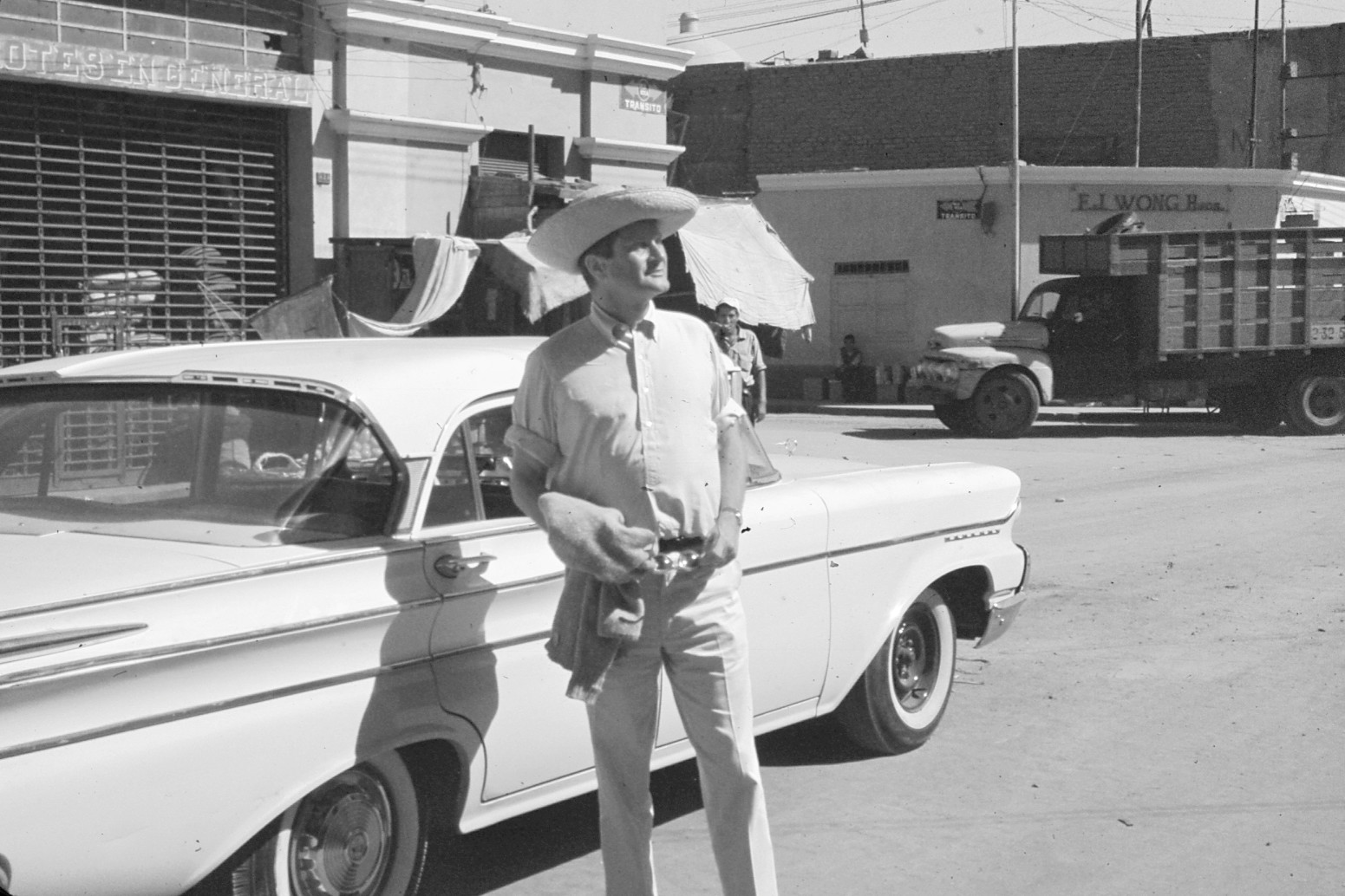 A black-and-white photograph of a man wearing a cowboy hat standing in front of a car on the street.