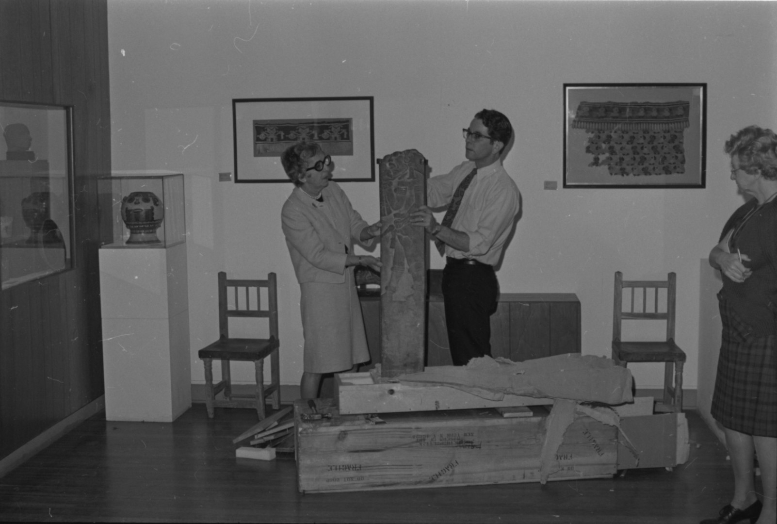 A black-and-white photograph of a man and woman holding up the Veracruz stone slab as they unpack it from a crate. Two framed textiles hang on the wall behind them, and a ceramic vessel is in a case to their right. On their left, at the very edge of the photograph, another woman watches.