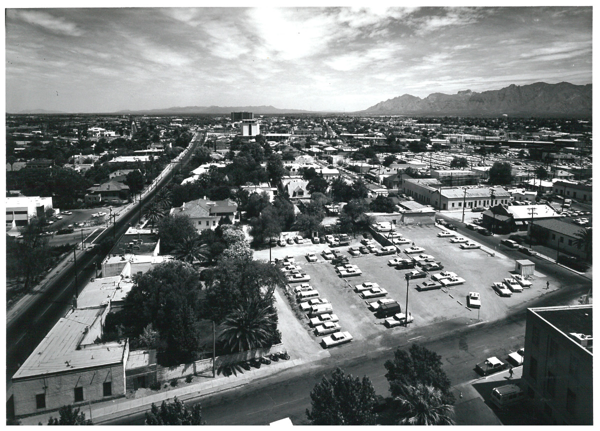 A black-and-white photograph of Tucson overlooking a parking lot and a number of buildings. The mountains can be seen in the far horizon.