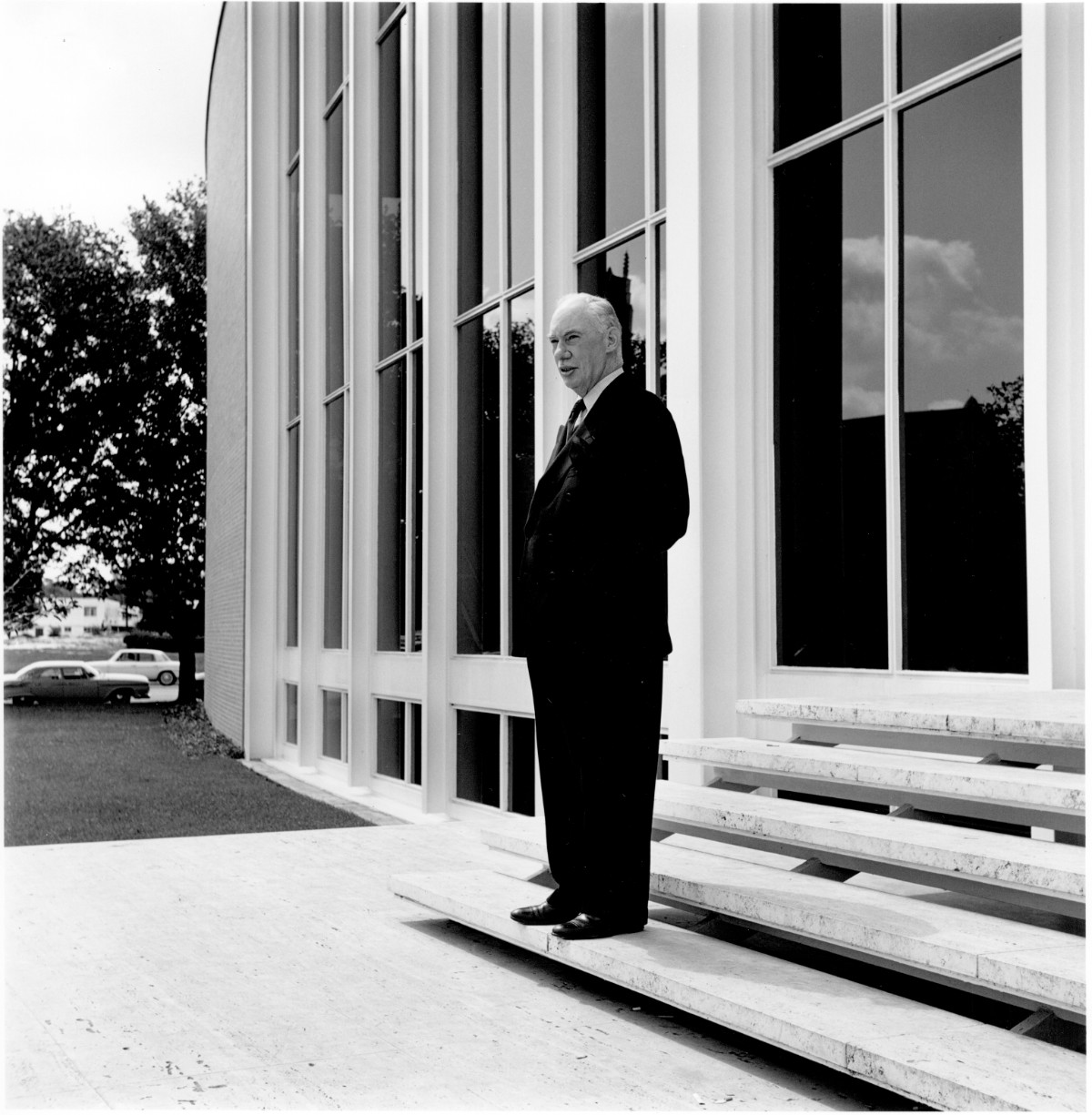 A black and white photograph of an older man dressed in a full suit stands on stone steps of a building with tall windows. The man looks off into the distance instead of at the camera.