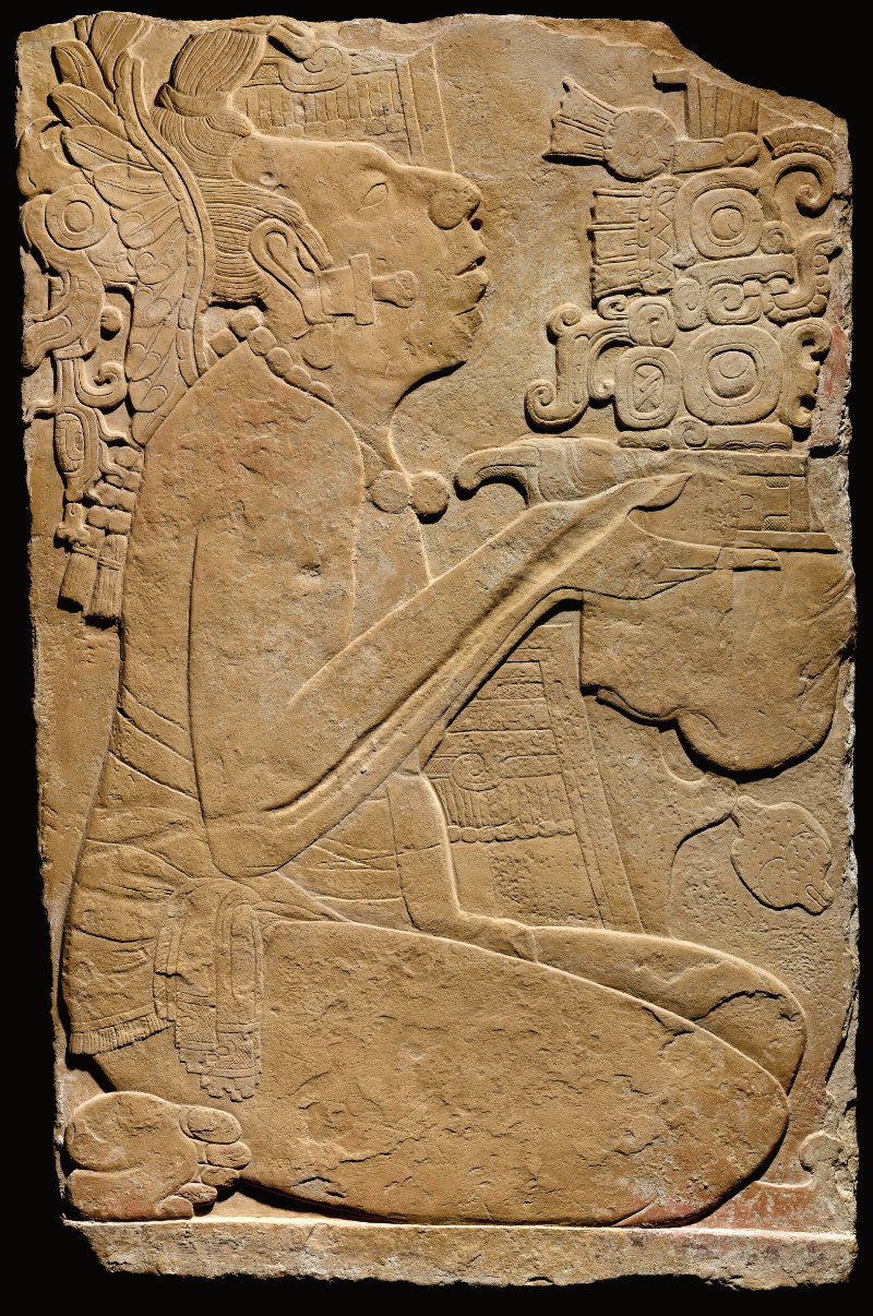 A bas relief panel carved out of stone of a figure seated on their knees with head tilted back, holding up an offering. The edges of the panel are worn but evenly cut.
