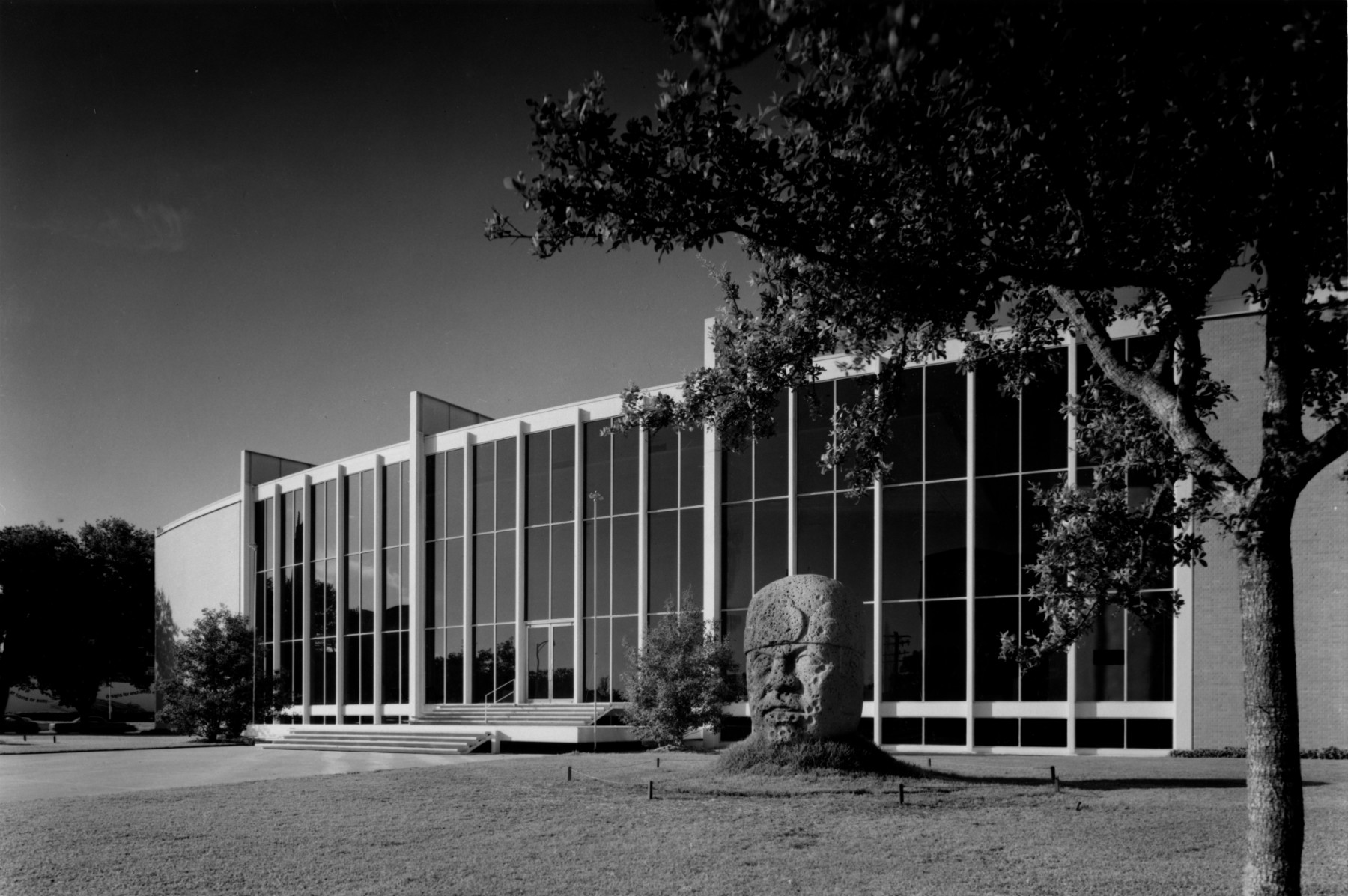 A black and white photograph from a distance of a building with large windows. There is an Olmec stone head placed in the grass next to the stairs leading into the building.