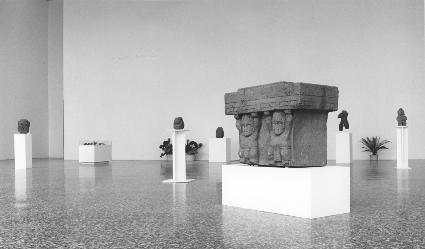 A black and white photograph of seven pedestals with stone sculptural objects on them in a large hall with polished stone floors and neutral painted walls.