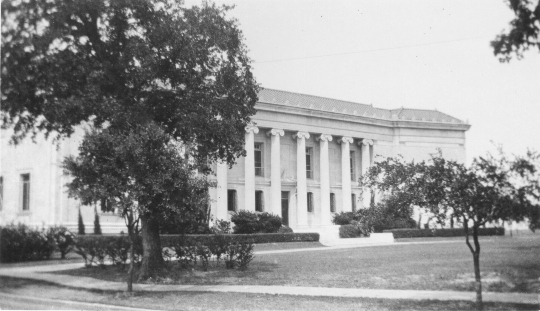 A black and white photograph of the front of a mansion with large columns in the entryway. A tall tree partially obscures the building.