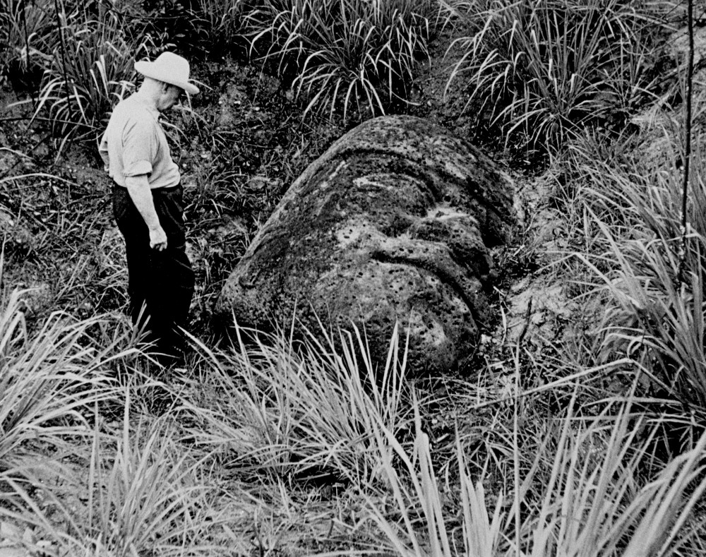 A black and white photograph of a man turned away from the camera facing an Olmec stone head half buried in the ground and overgrown by plants.