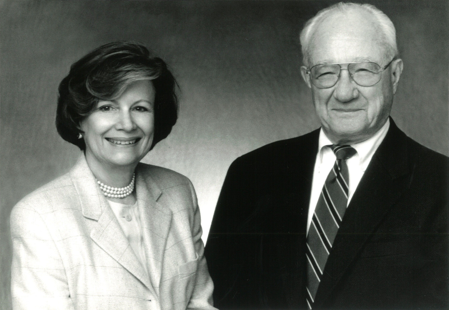 A black-and-white photograph of a man and woman seated in front of a neutral background. The woman wears a light-colored suit, a pearl necklace, and her hair is short. The man wears a dark colored suit, striped tie, and glasses. They are both smiling.
