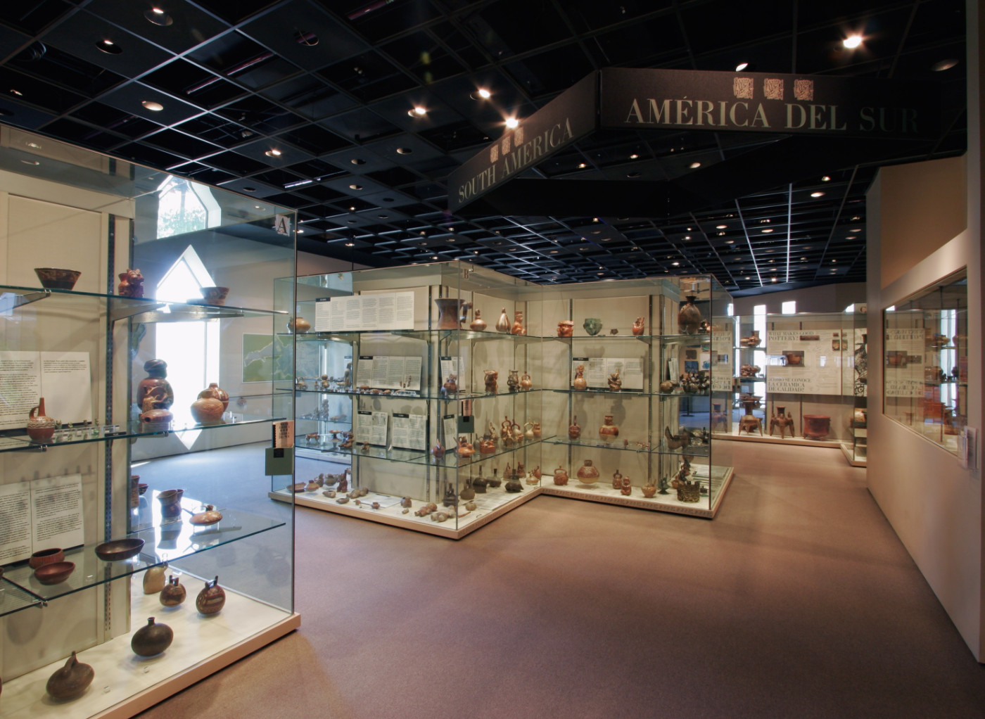 A color photograph of a gallery with carpeted floor and diamond shaped windows. A large sign hanging overhead reads, South America. The cases are large and contain multiple shelves, didactic text panels, and large numbers objects.