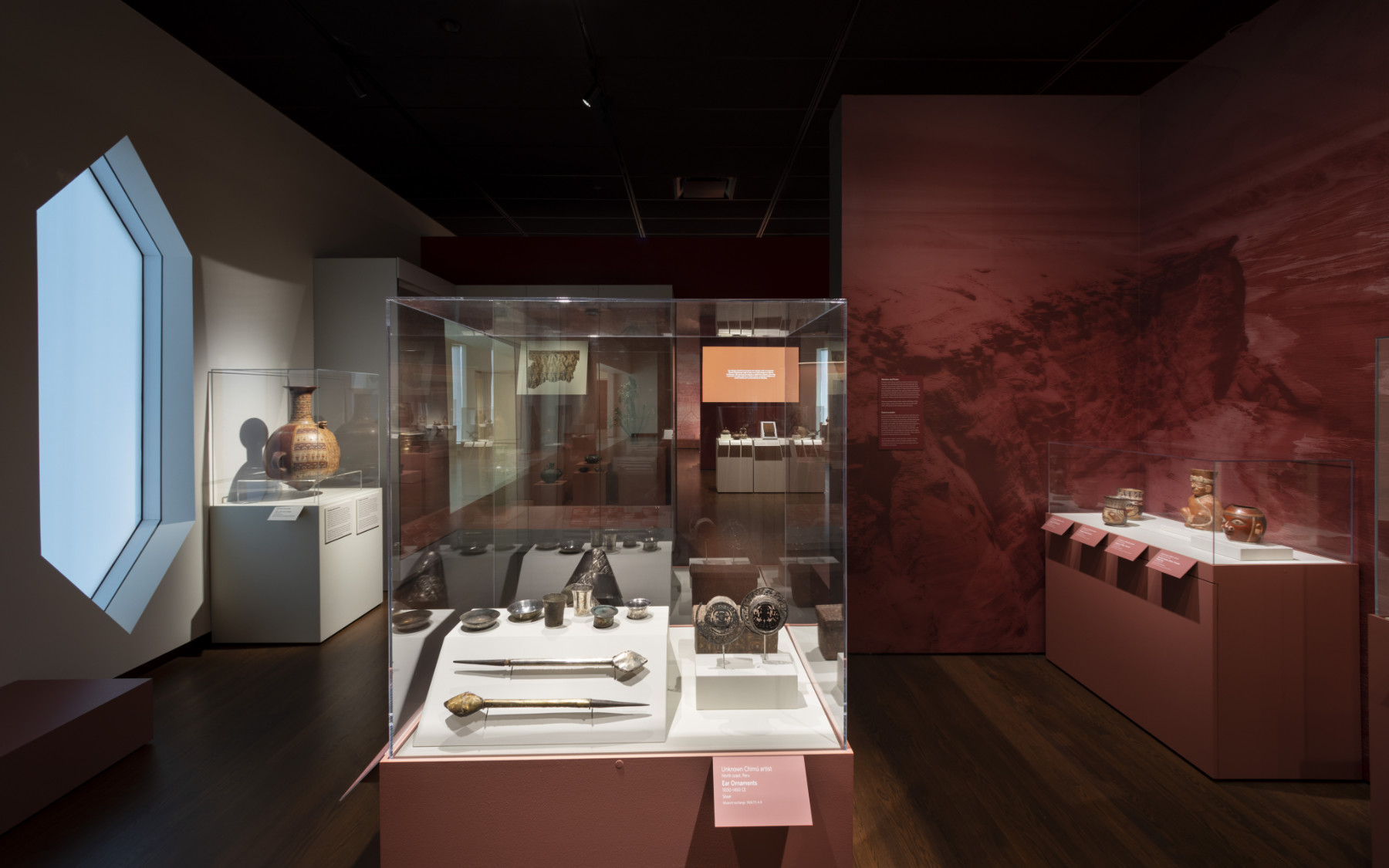 A color photograph of a gallery displaying ancient American artworks with hardwood floor and diamond shaped windows. The cases and walls are painted red.