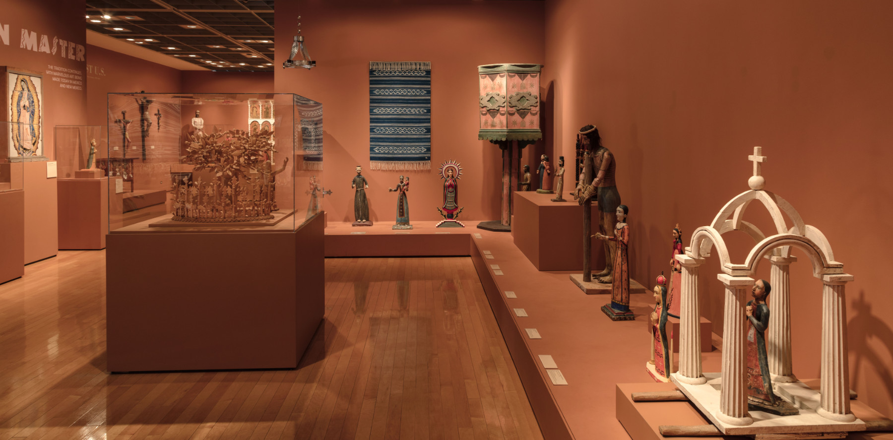 A color photograph of a gallery painted orange displaying polychromatic wooden religious objects, textiles, and metalwork.