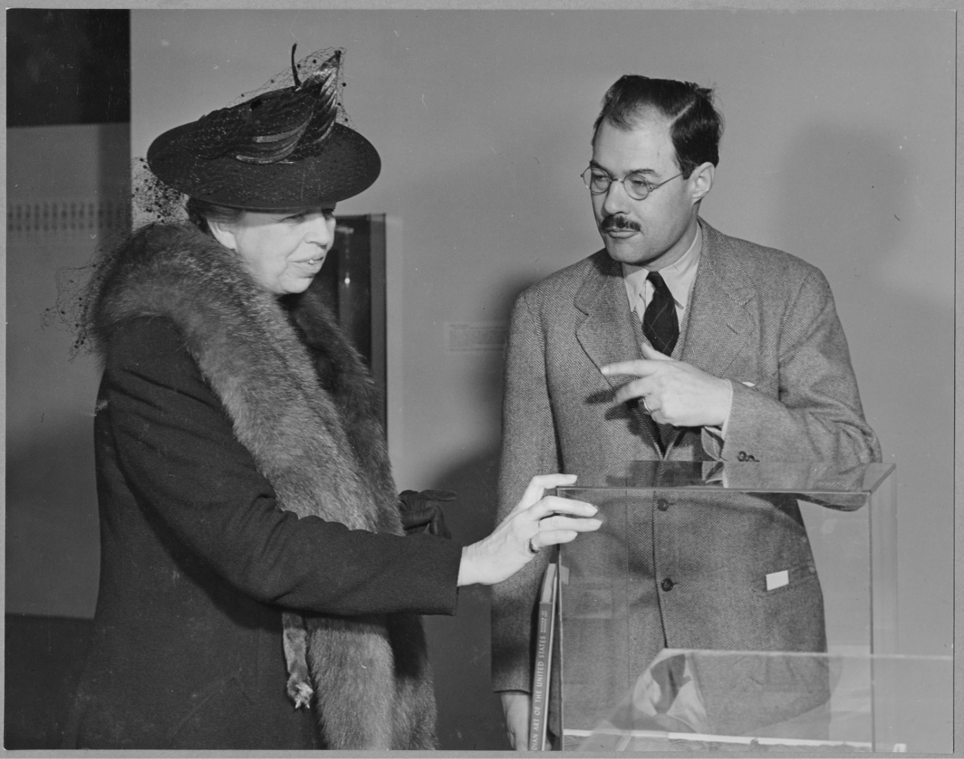 A black-and-white photograph of a man and woman talking in an exhibition. The woman wears a fur shawl and a dark hat. The man wears a suit and glasses and has a mustache.