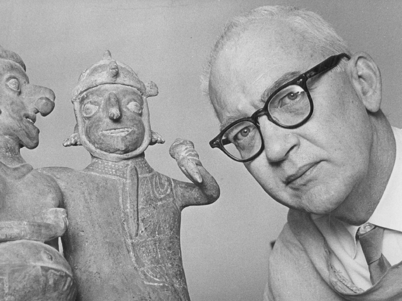 A black-and-white photograph of an older man leaning in next to two ceramic figurines. He is seen from the neck up and wears dark glasses.