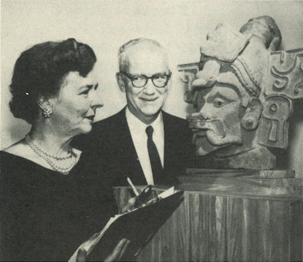 A black-and-white photograph of a man and woman standing in front of a ceramic figure in the shape of a human head and headdress. The man looks at the woman, and the woman holds a clipboard and pen.