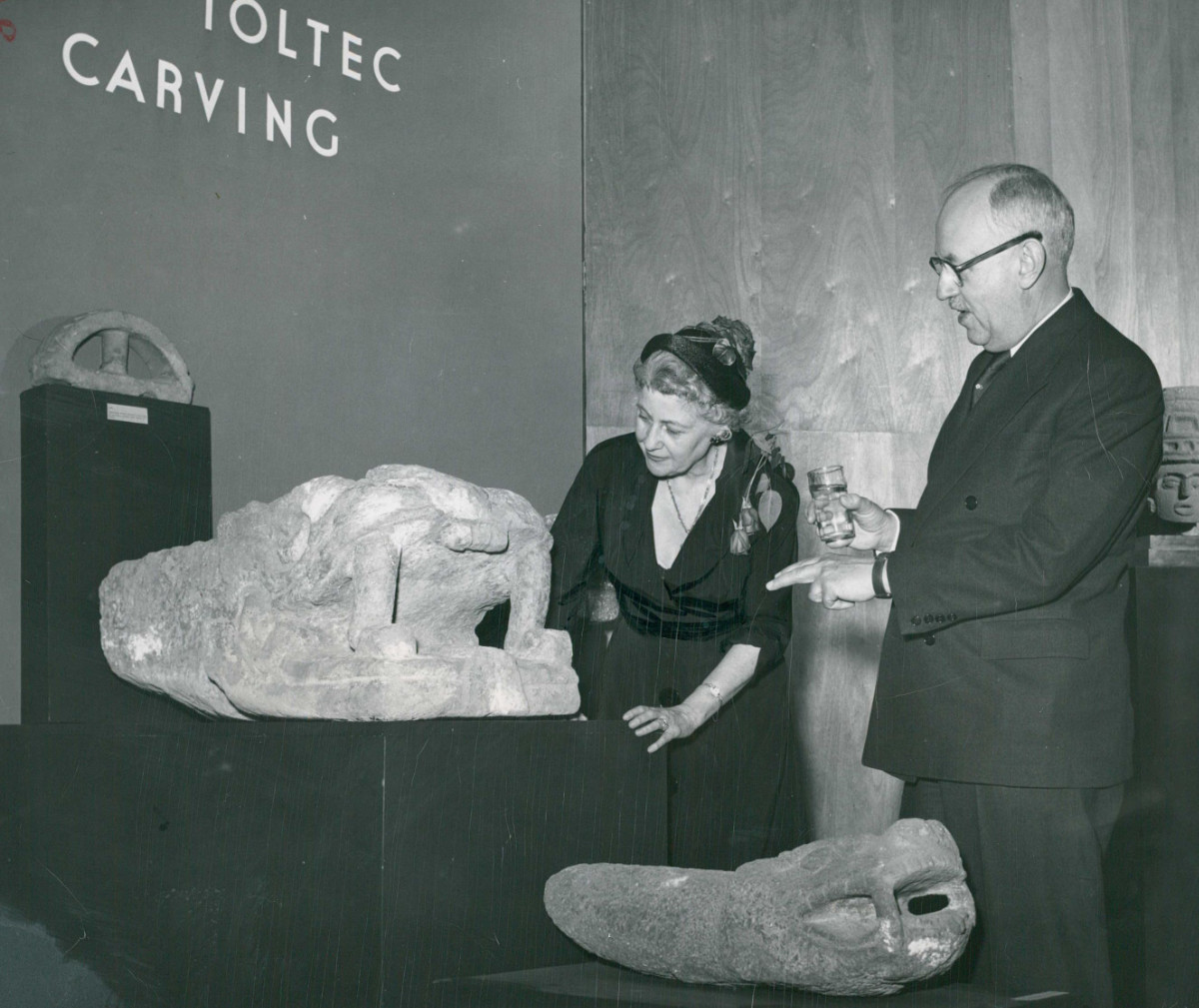 A black-and-white photograph of a man and woman looking at a stone sculpture of a serpent head. The woman leans over it, and the man with a drink in hand points at the artwork. On the wall, above the artwork, reads, “Toltec Carving.”