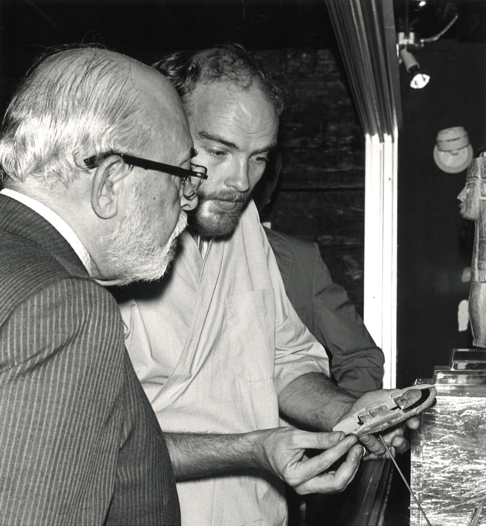 A black-and-white photograph of two men looking at a stone sculpture. The man to the left is wearing a pinstripe suit and glasses while the man to the right holds the object they are looking at.