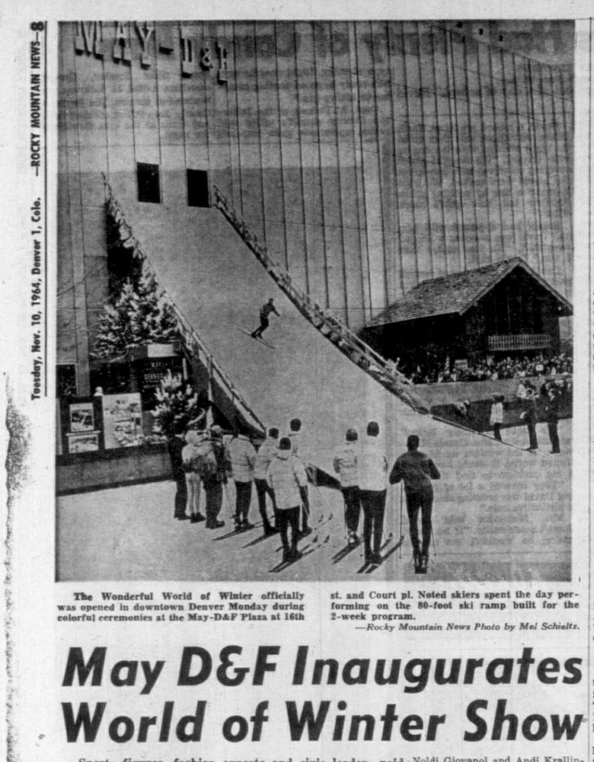 A newspaper clipping from the Rocky Mountain News dated November 10, 1964. The headline below the image reads, May D&F Inaugurates World of Winter Show. The photograph depicts a ski slope attached to the outside of a building with a crowd of people watching a skier descend. The caption reads, The wonderful world of winter officially was opened in downtown Denver Monday during colorful ceremonies at the May-D&F Plaza at 16th st. and Court pl. Noted skiers spent the day performing on the 80-foot ski ramp built for the 2-week program.