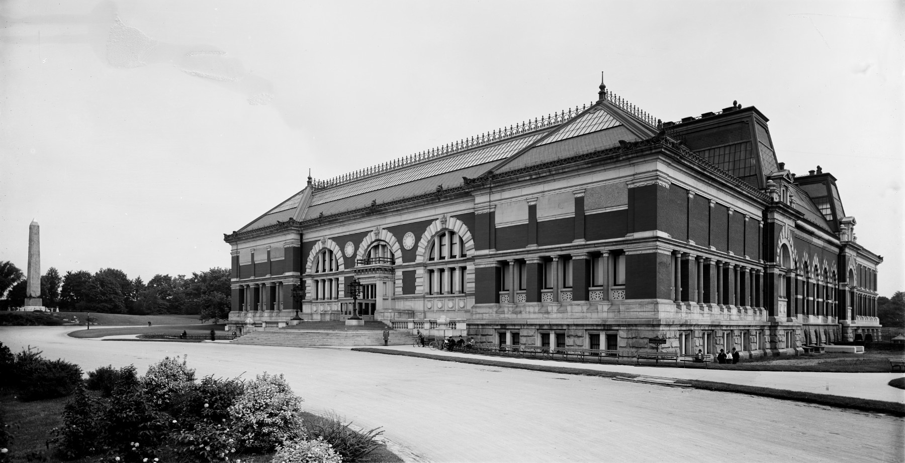 A black-and-white photograph in three-quarter view of a large brick building. The building has a gabled roof with wrought-iron trim and arched double-height windows with stone trim at the grand entryway.