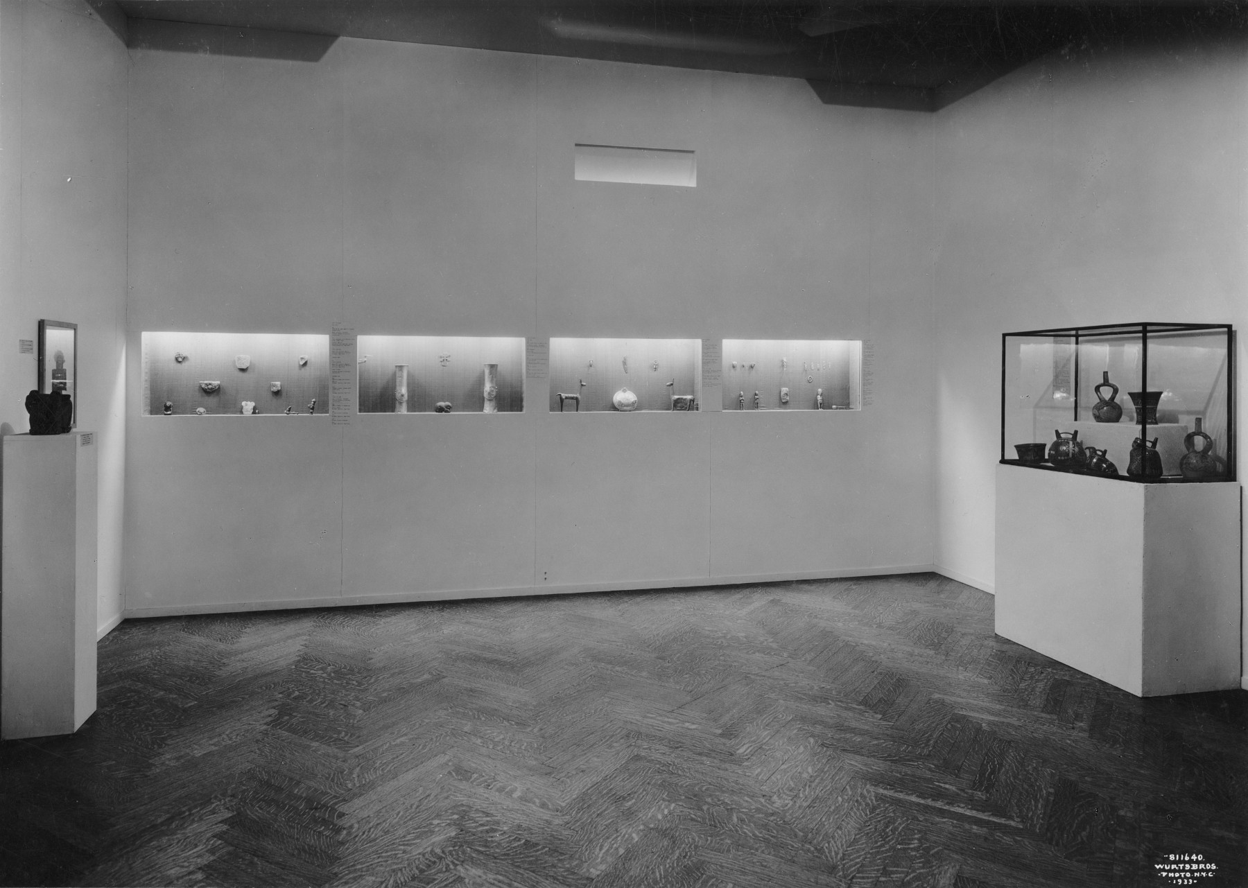 A black-and-white photograph of a gallery space with one pedestal and one case displaying artwork on either side of the room, facing each other. The back wall has four vitrine openings to showcase more artwork.