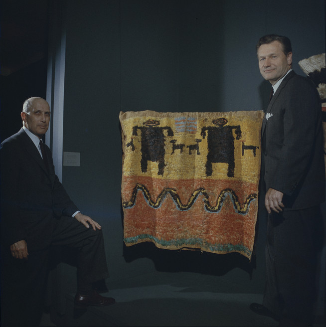 A color photograph of two men in formal wear posed in front of a yellow-and-orange feather mantle depicting dark figures and animals above a curved line.