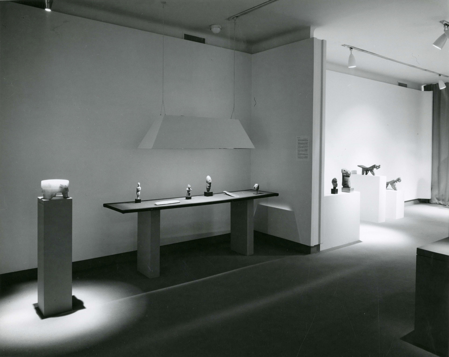 A black-and-white photograph of two adjacent gallery spaces separated by a half wall. Each room contains pedestals and tables displaying ceramic and stone works that are spotlit from above.
