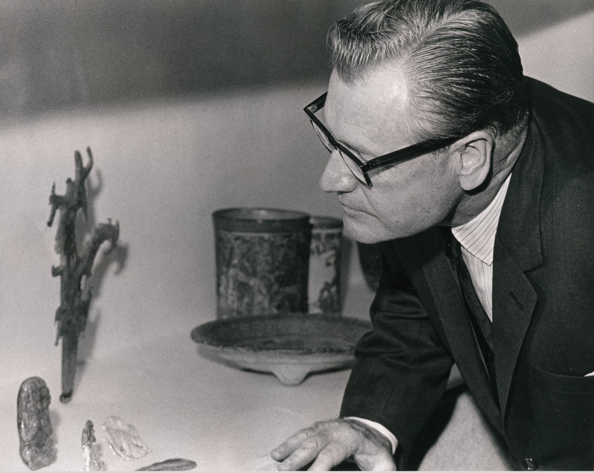 A black-and-white photograph of a man in formal wear and glasses, looking closely at four small stone sculptures displayed near two ceramic vessels and a plate.