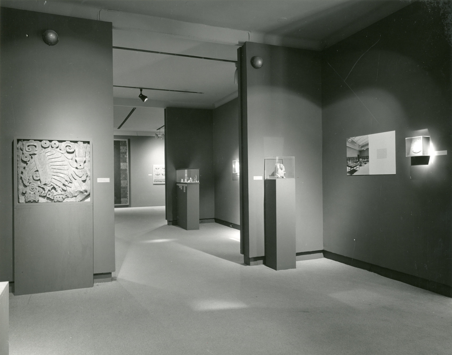 A black-and-white photograph of three adjacent gallery spaces with dark-colored walls and pedestals that contain a few stone, ceramic, and textile works.
