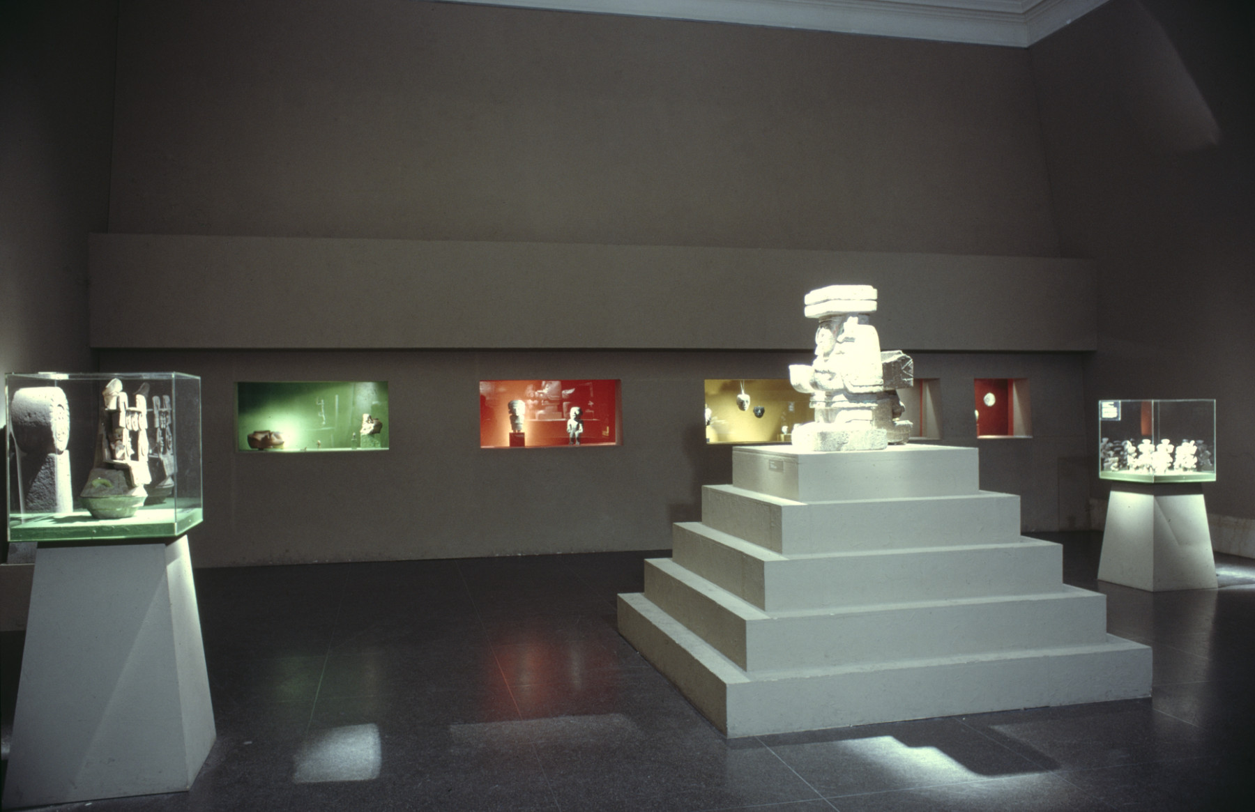 A color photograph of a gallery where the back wall contains five colorfully lit vitrines, containing stone and ceramics artworks. A pyramid-shaped pedestal, bearing a stone sculpture on top, is in the center, with two smaller cases off to the side.