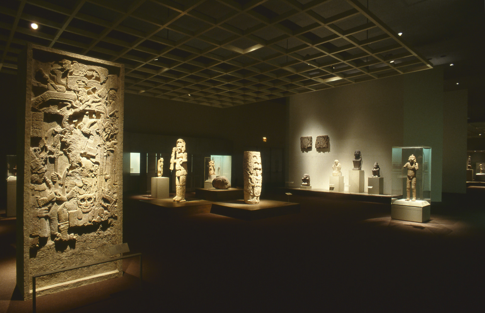 A color photograph of a gallery with a large stone stela in the left foreground with other stone sculptures on pedestals and cases dispersed throughout the space.  The space is dimly lit with spotlights on the artworks.