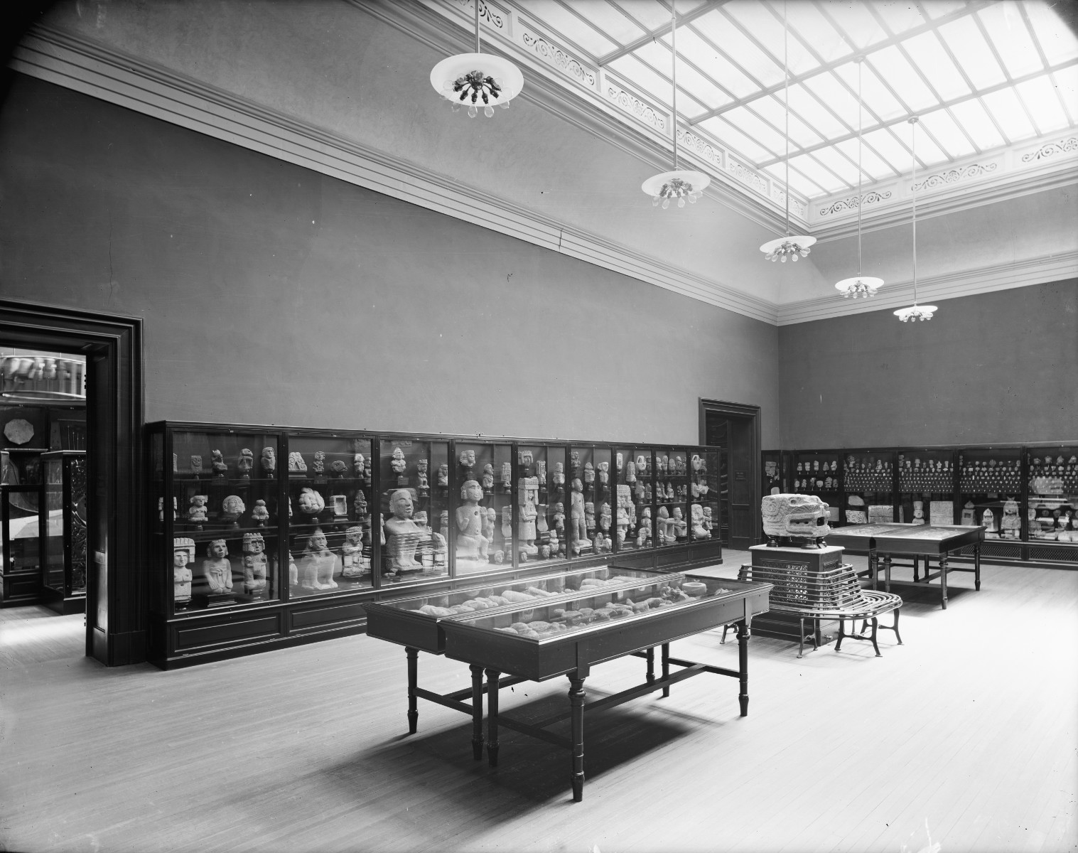 A black-and-white photograph of a gallery with hardwood floors and display cases lining the walls and in the center of the space, where various ancient American objects can be seen. Five chandeliers hang above from the skylight.