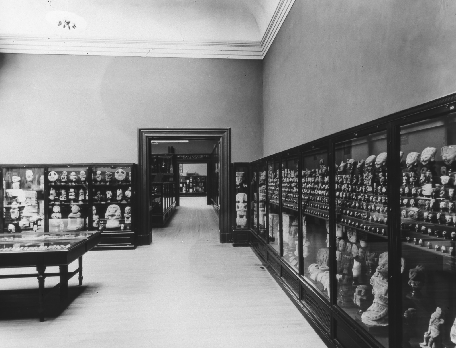 A black-and-white photograph of a gallery with hardwood floors and display cases lining the walls and in the center of the space. Through an open doorway another gallery is visible.