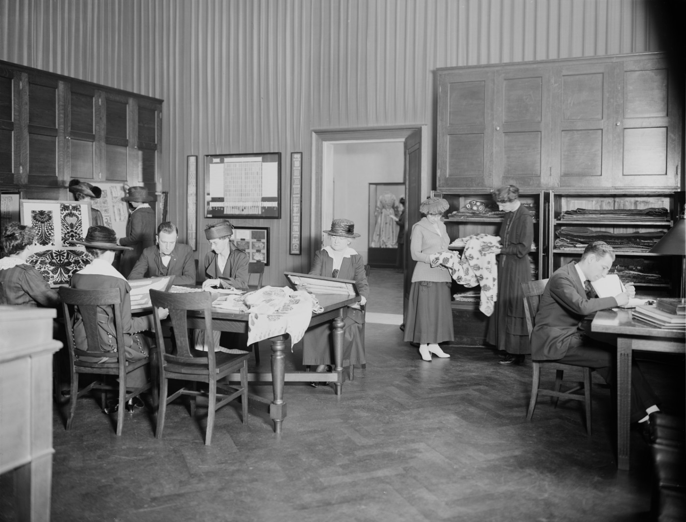 A black-and-white photograph of a study room, where people stand near cabinets and sit at tables, studying various textiles.