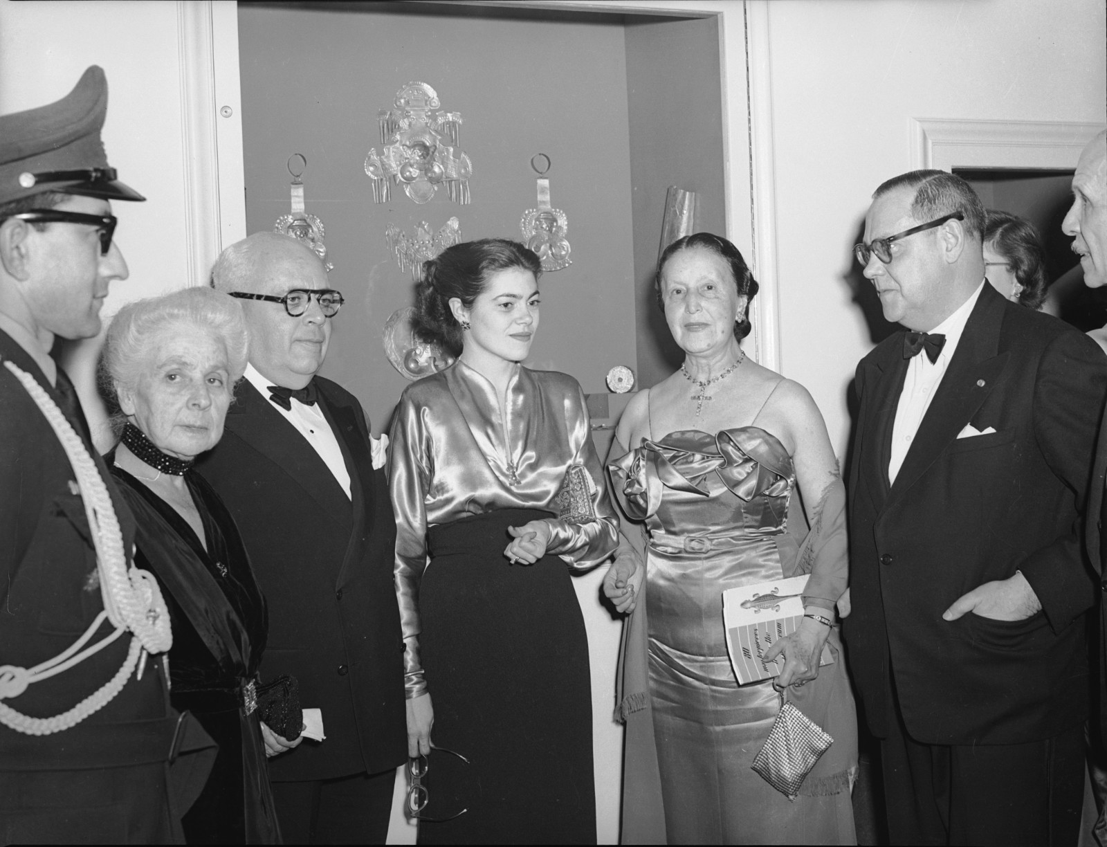 A black-and-white photograph of a group of men and women in formal wear standing together in front of a display case featuring gold objects.