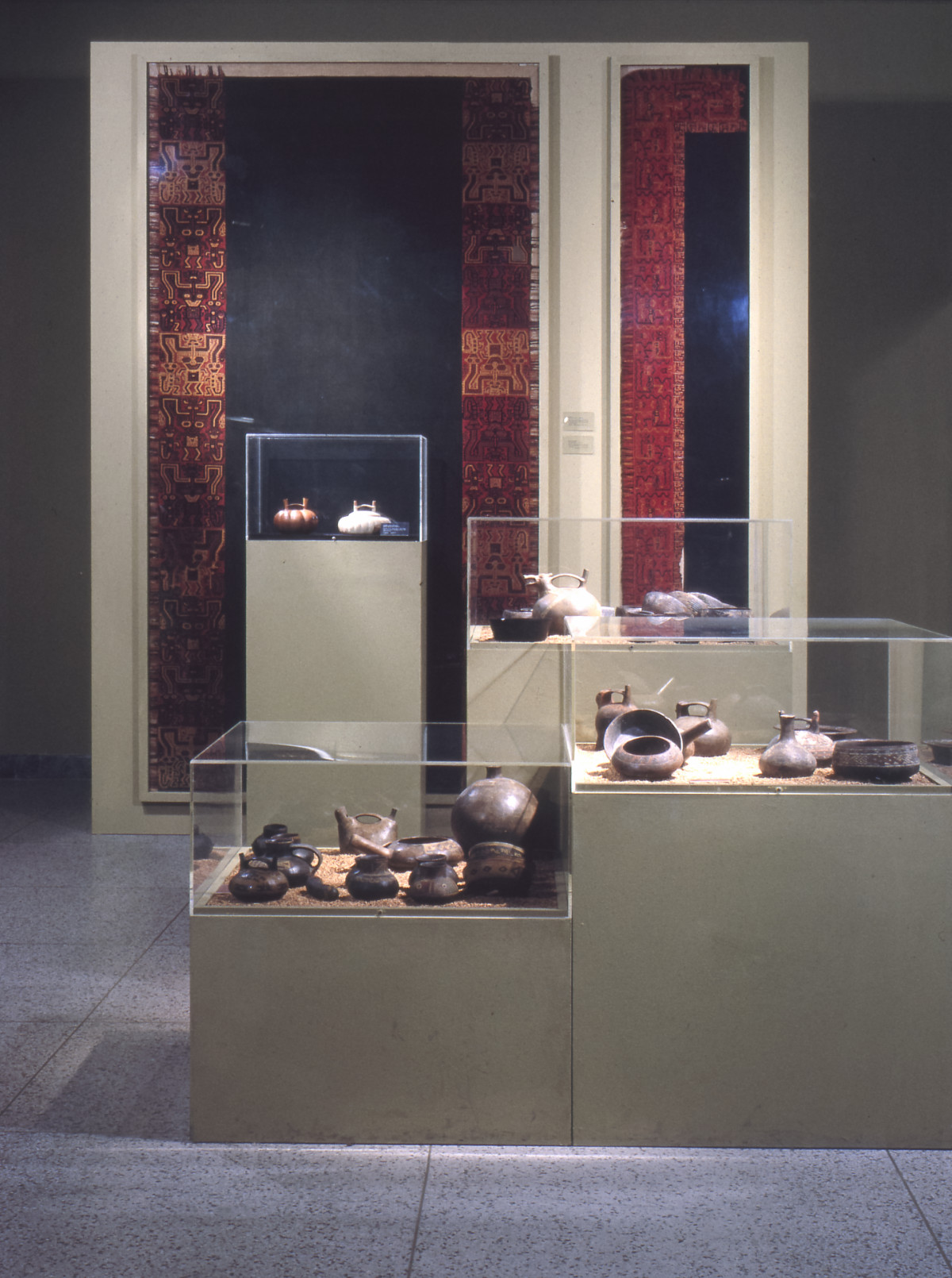 A color photograph of a gallery with four display cases of varying heights, each containing many dark brown ceramic double-spouted vessels. Behind the cases, two large black and red textiles are framed on the wall.